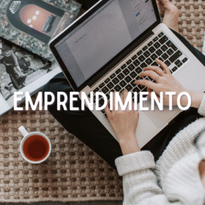 Persona trabajando en una laptop colocada sobre sus piernas, sentada en el suelo sobre una alfombra. A un lado hay una taza con bebida caliente y una revista abierta. Sobre la imagen aparece la palabra ‘EMPRENDIMIENTO