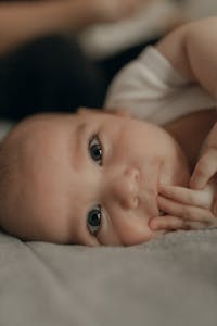 Adorable baby with big eyes lying down indoors, touching lips with fingers, embodying curiosity and innocence.