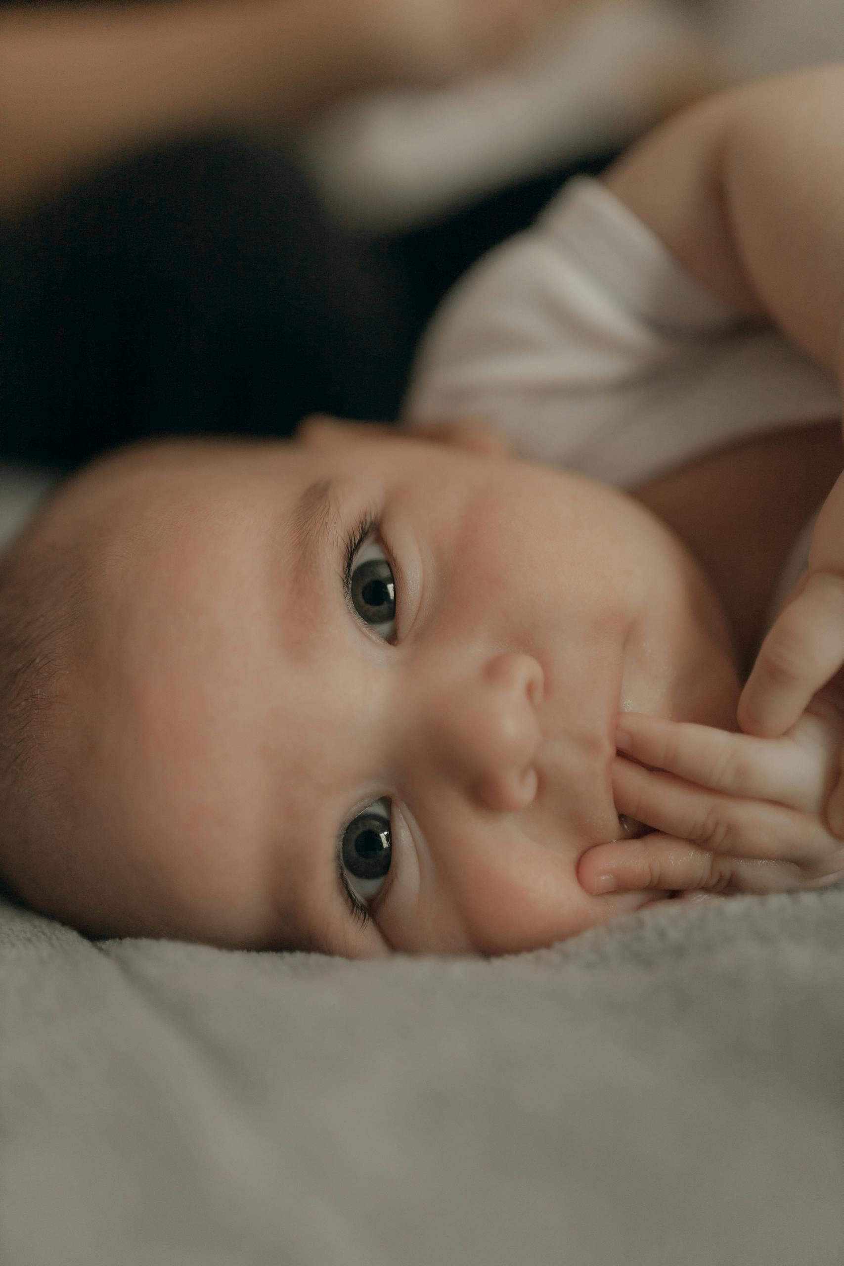Adorable baby with big eyes lying down indoors, touching lips with fingers, embodying curiosity and innocence.