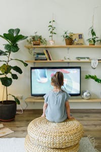 Back view of unrecognizable little kid watching television in living room with potted plants in house