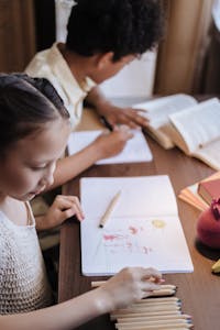 Children focused on drawing and studying at a desk with books and colored pencils.