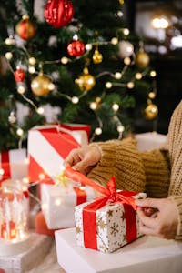 Crop anonymous female in warm sweater unpacking gift box decorated with red ribbon bow while sitting near Christmas tree during holidays