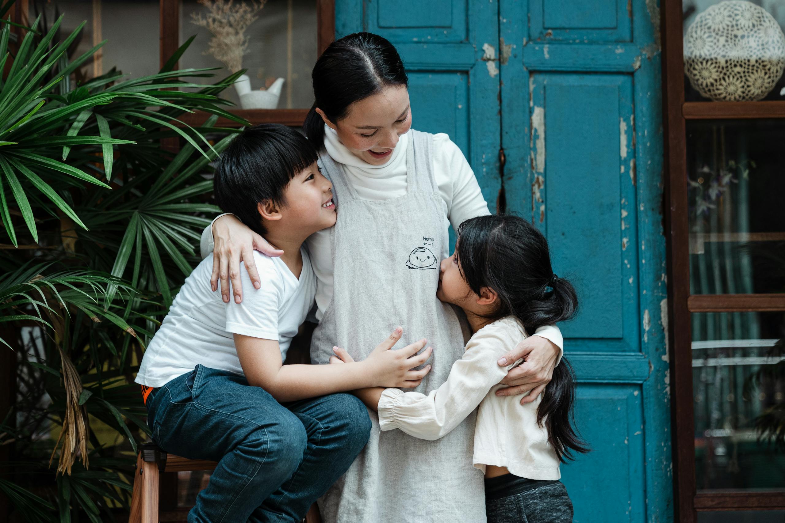 Happy smiling Asian woman with daughter and son hugging against aged building and plant on daytime